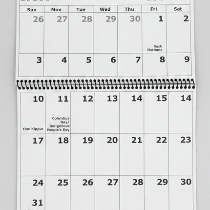 A spiral-bound October 2027 desk calendar page showing the dates and day names, photographed from above on a dark wood surface.