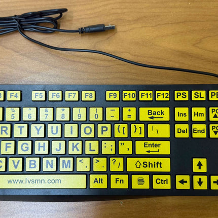 A black computer keyboard with bright yellow function keys and a black cable, placed on a wooden desk.