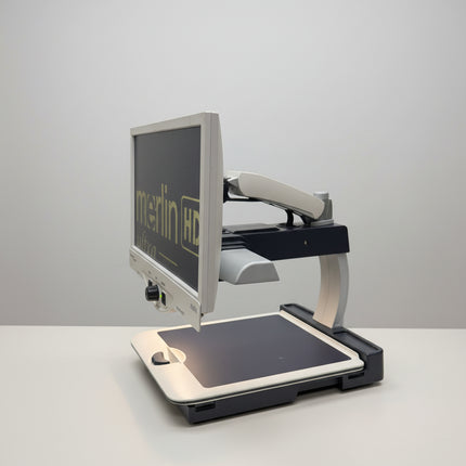 A desktop monitor and keyboard setup on a metal cart in a warehouse or office space, with shelving and boxes visible in the background. The monitor displays the text 'merlin HD'.