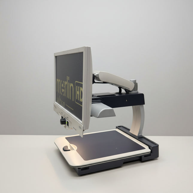 A desktop monitor and keyboard setup on a metal cart in a warehouse or office space, with shelving and boxes visible in the background. The monitor displays the text 'merlin HD'.