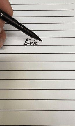 A hand holding a bold writing pen for low vision, writing the name 'Eric' on lined paper.