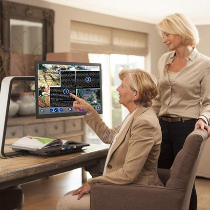 Woman using a desktop video magnifier for low vision with a support person in a bright home environment.