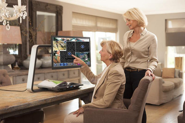 Woman using a desktop video magnifier for low vision with a support person in a bright home environment.