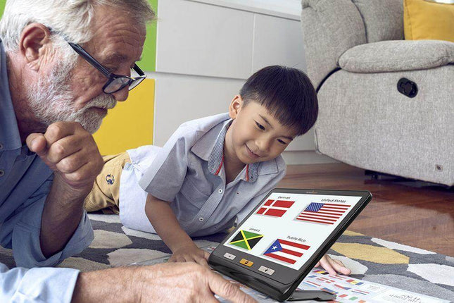 A senior man and a child using a portable video magnifier for low vision to read country flags on a colorful rug.