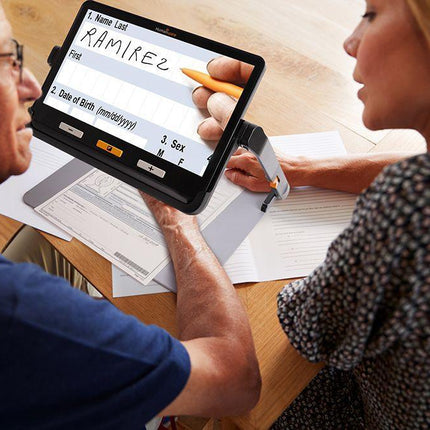 Portable video magnifier for low vision in use by two people at a table, showcasing clear text on the screen.