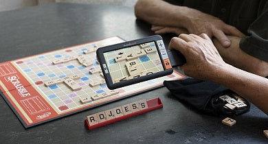 Person using a handheld electronic video magnifier to read a Scrabble game board.