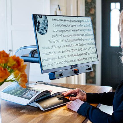 Person using a 24 inch video magnifier for low vision to read a book at a table with flowers.