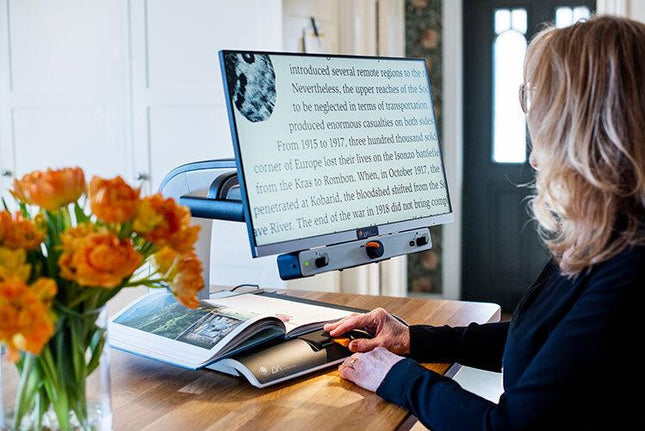 Person using a 24 inch video magnifier for low vision to read a book at a table with flowers.