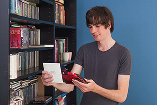 User utilizing a used video magnifier to read a card in front of a bookshelf filled with books and CDs.