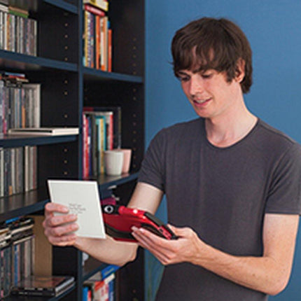 Man using a RUBY 7" HD portable video magnifier to read a card while standing near bookshelves.