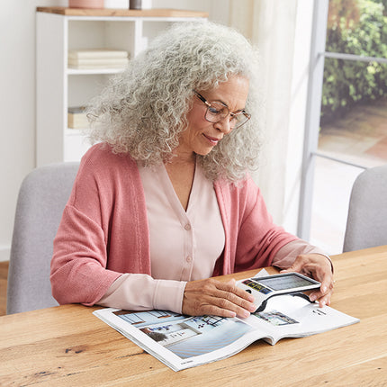 An older woman using Smartlux Digital, a portable HD video magnifier for low vision, while reading a magazine.