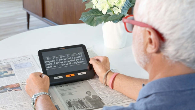 A user reading a newspaper with the Explore 8 handheld electronic magnifier on a table.