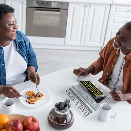 Two people enjoying breakfast together, using a handheld electronic magnifier on a table with coffee and fruits.