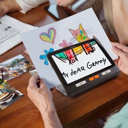 Person using a handheld electronic magnifier to read a colorful drawing while sitting at a table.